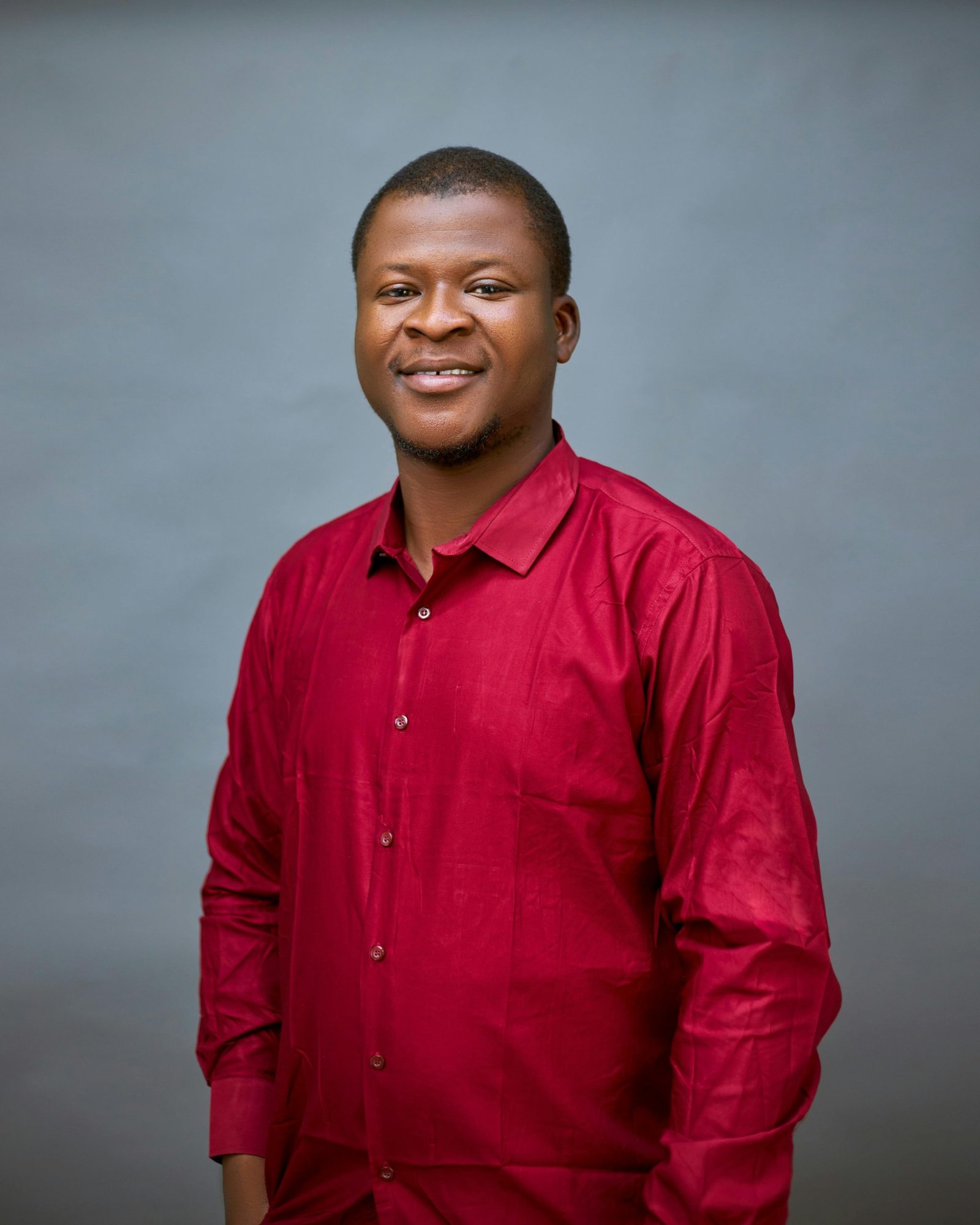 Portrait of a smiling African adult male in a red shirt against a plain background.